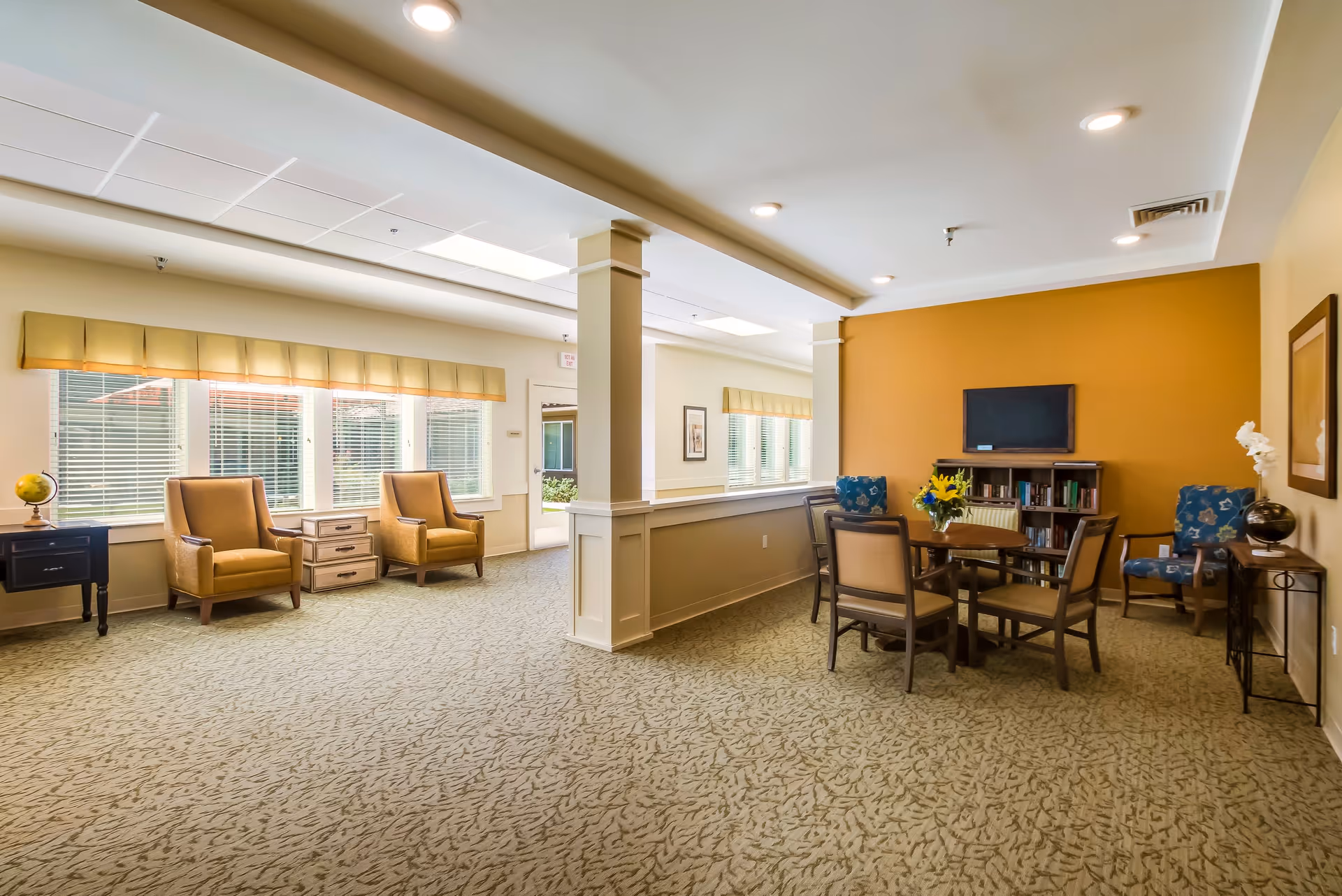 A bright and spacious common area in a senior living facility with beige carpet and walls. The room features two tan armchairs near large windows with blinds and a valance, a round wooden table with four chairs, a small bookshelf with a flat-screen TV mounted above it, and two blue patterned armchairs. There are decorative items including a globe, a vase with flowers, and framed artwork on the walls.