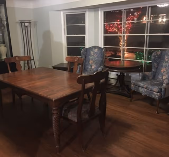 A dining room with a wooden dining table surrounded by four wooden chairs with cushioned seats. In the background, there are two upholstered armchairs with floral patterns and a round wooden side table between them. Large windows show outdoor trees decorated with red and green lights at night.