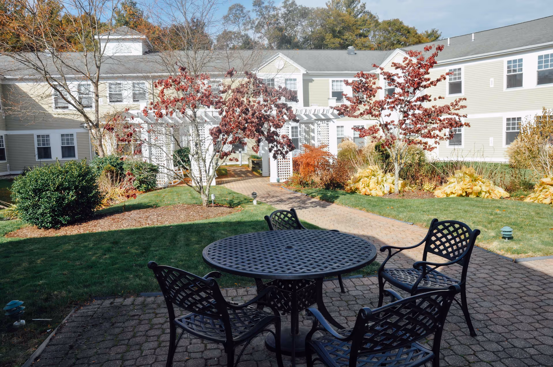 Outdoor patio area with a round metal table and four matching chairs on a brick-paved surface. The patio overlooks a landscaped garden with green grass, bushes, and trees with red and orange autumn leaves. In the background, there is a two-story light-colored building with many windows and a white pergola structure.