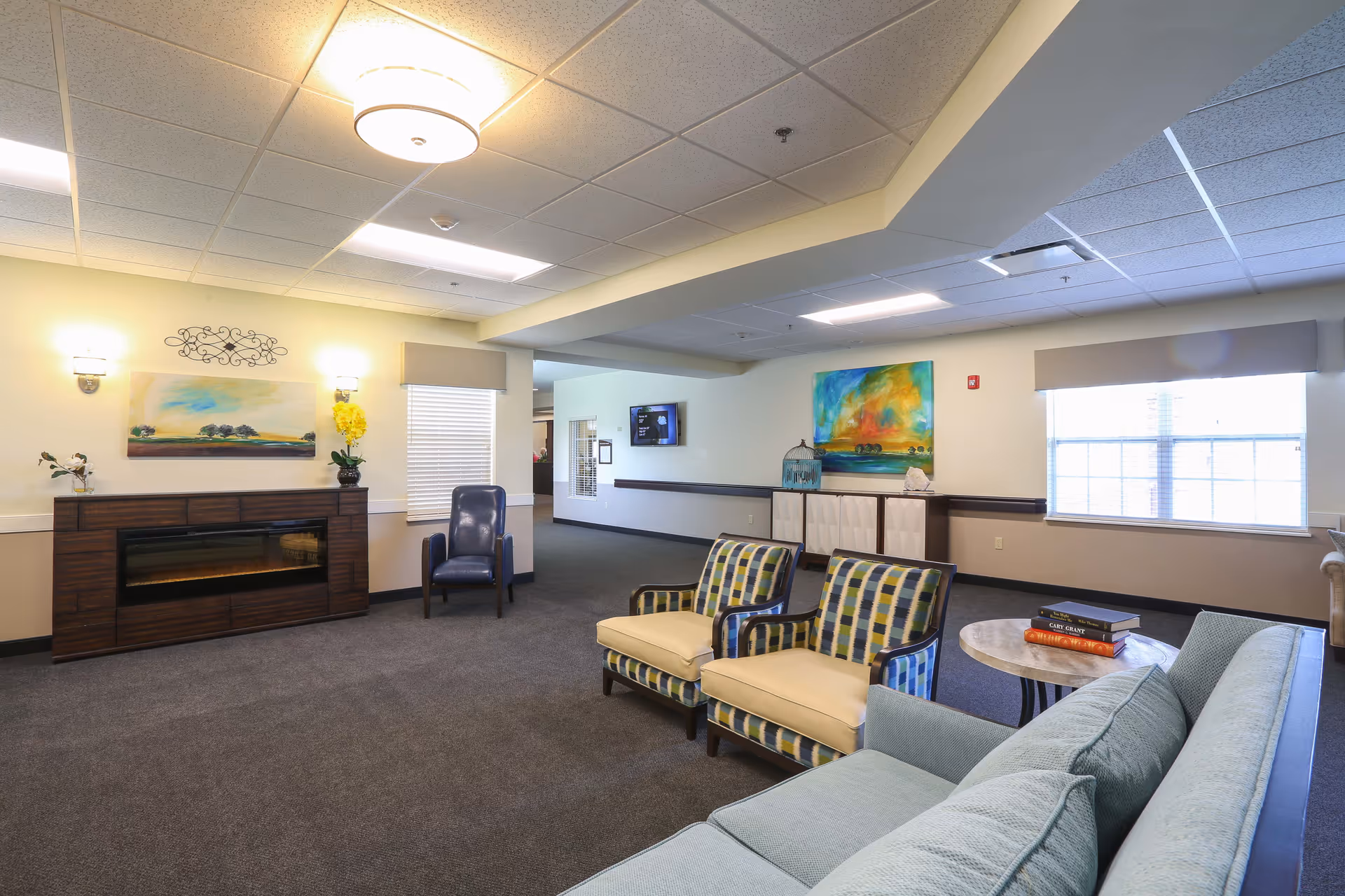 A spacious senior living facility common area with a light blue sofa, two patterned armchairs, a round table with books, a dark blue chair near a window, and a wooden electric fireplace with a landscape painting above it. The room has beige walls, carpeted floor, ceiling lights, and large windows letting in natural light.