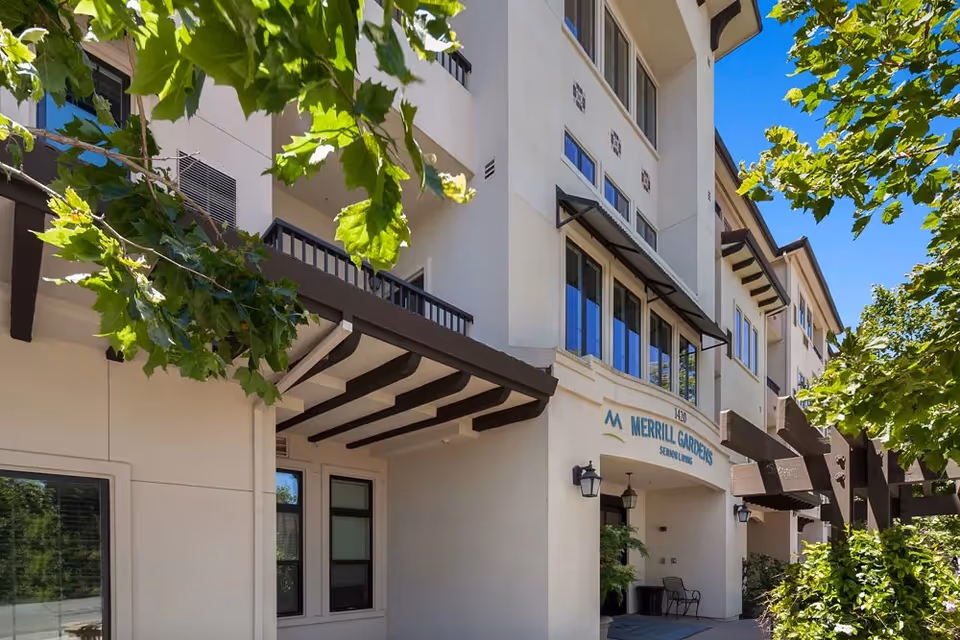 Exterior view of Merrill Gardens at Willow Glen senior living facility showing a multi-story building with balconies, large windows, and a covered entrance. Green leafy trees partially frame the building under a clear blue sky.
