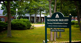 Exterior view of Windsor Skilled Nursing-Rehabilitation Center with a green sign and trees.