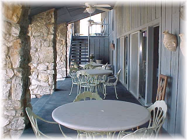 Covered outdoor patio area with round tables and chairs arranged along a stone wall and a wooden paneled wall with windows. Ceiling fans are mounted overhead and a staircase is visible at the far end.