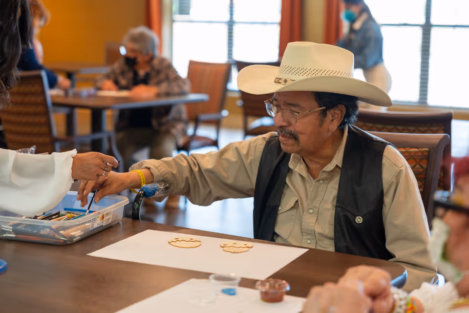 An older man wearing a cowboy hat paints wooden shapes at a table during a group activity in a communal room.