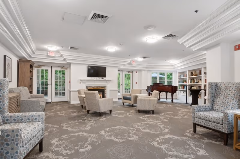 A spacious and well-lit living room area in a senior living facility featuring multiple upholstered armchairs and sofas arranged around a fireplace with a mounted flat-screen TV above it. The room has large windows and glass doors allowing natural light to enter, a grand piano in the corner, built-in bookshelves filled with books and decorative items, and patterned carpet flooring.