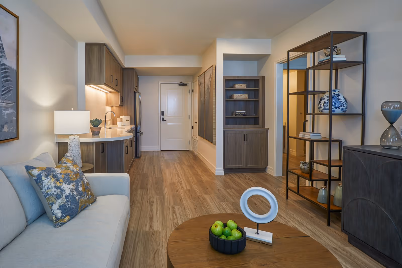 Well-lit living area and kitchenette featuring a sofa with a patterned pillow, round coffee table with fruit, open shelving, and an entry door down a wood-floor hallway.