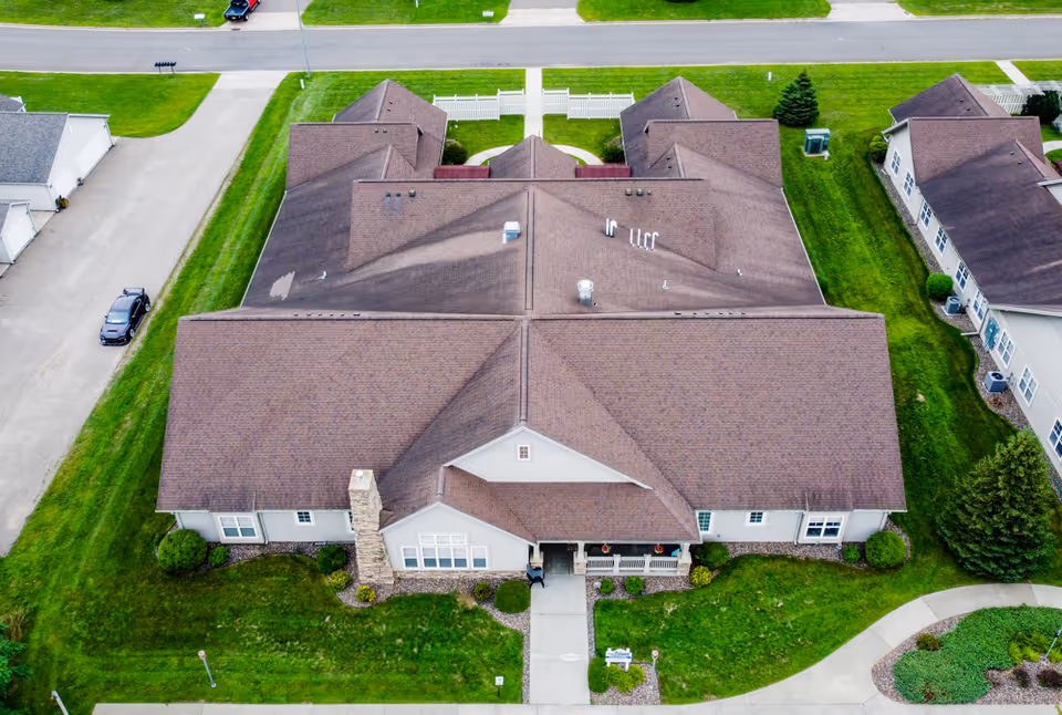 Aerial view of a single-story senior living facility building with a brown roof, surrounded by green lawns and neighboring buildings. The building has a front entrance with a small porch and a walkway leading to the sidewalk.