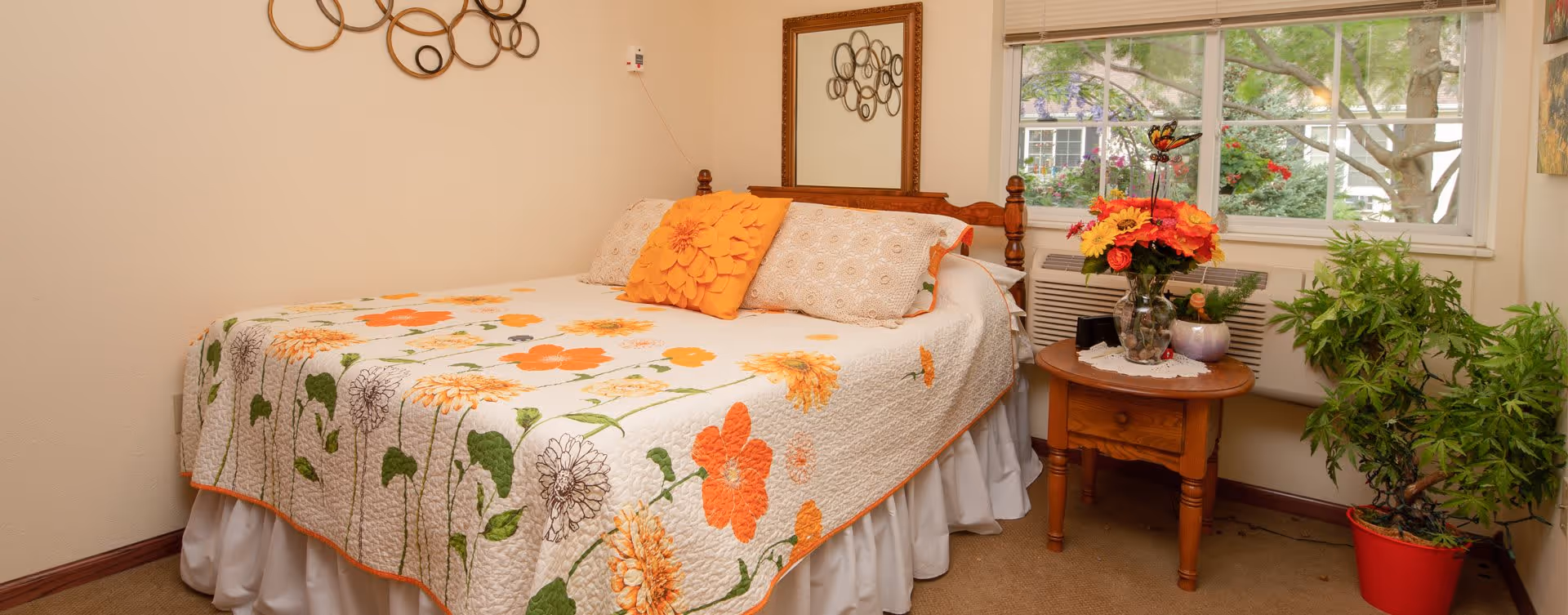 A cozy bedroom with a bed covered in a floral quilt featuring orange and yellow flowers. The bed has decorative pillows, including one with a large orange flower design. Next to the bed is a wooden nightstand with a vase of colorful flowers and a small potted plant. A large window behind the nightstand shows greenery outside. The room also has a decorative wall mirror and a circular metal wall art piece.