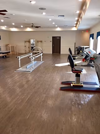 A spacious rehabilitation or physical therapy room with wooden flooring, parallel bars for walking exercises, exercise equipment including a seated pedal machine, ceiling fans, and bright overhead lighting.