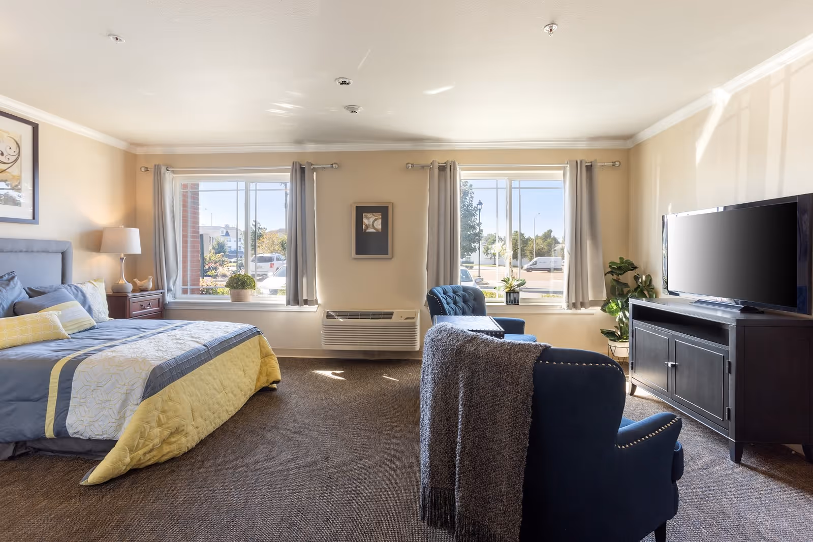 A bright and spacious bedroom in an assisted living facility featuring a large bed with gray and yellow bedding, a nightstand with a lamp, two large windows with gray curtains, a blue armchair with a gray throw blanket, a dark wood TV stand with a flat-screen television, and some potted plants near the windows.