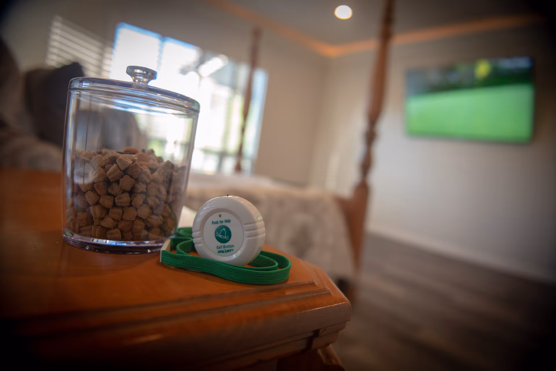 A close-up of a wooden bedside table with a glass jar filled with brown pet treats and a white medical alert call button with a green lanyard. In the background, there is a blurred view of a bed with a wooden post and a wall-mounted television.