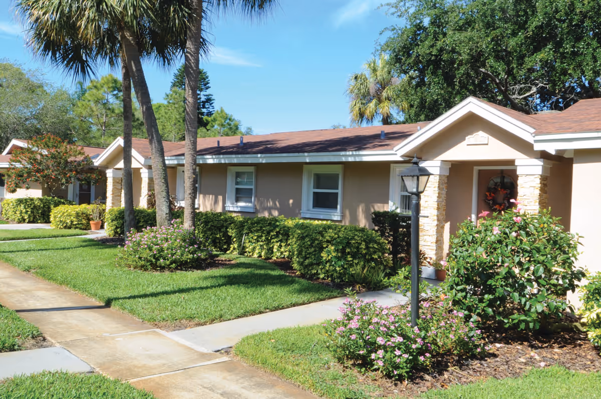 Exterior view of a single-story residential building with beige walls and a brown roof, surrounded by well-maintained green lawns, bushes, palm trees, and flowering plants under a clear blue sky.