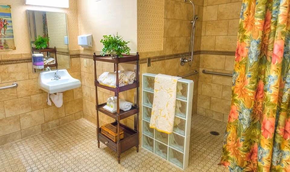 A bathroom with beige tiled walls and floor, featuring a wall-mounted white sink with a mirror above it. There is a wooden shelving unit holding towels, toiletries, and a potted plant. The shower area has a glass block half-wall and a floral shower curtain with orange, pink, blue, and green colors. Grab bars are installed near the sink and in the shower area.
