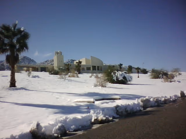 Exterior view of Hi-Desert Continuing Care Center building with snow-covered ground, palm trees, and clear blue sky.