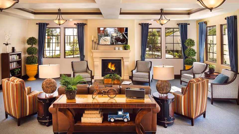 A cozy living room with a fireplace, surrounded by several armchairs and sofas. The room features large windows with blue curtains, potted plants, a wooden coffee table with decorative items, and warm lighting from ceiling fixtures and table lamps.