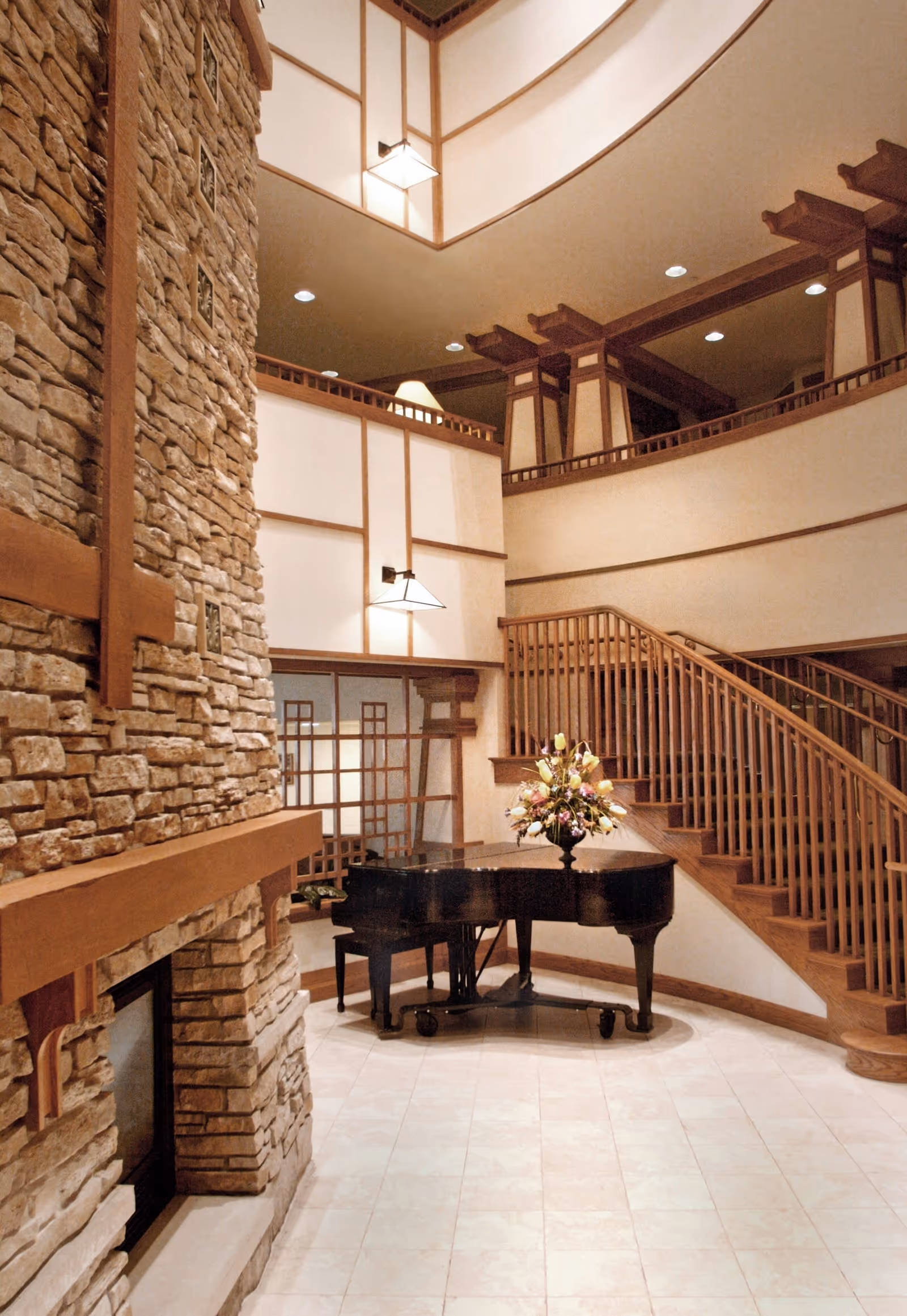 A warm atrium-style interior featuring a grand piano topped with flowers, a large stone fireplace, and a wooden staircase leading to upper balconies.