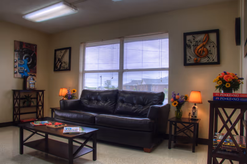 A cozy common room with a black leather sofa, coffee table, side tables with lamps and flowers, and framed wall art beneath a large window.