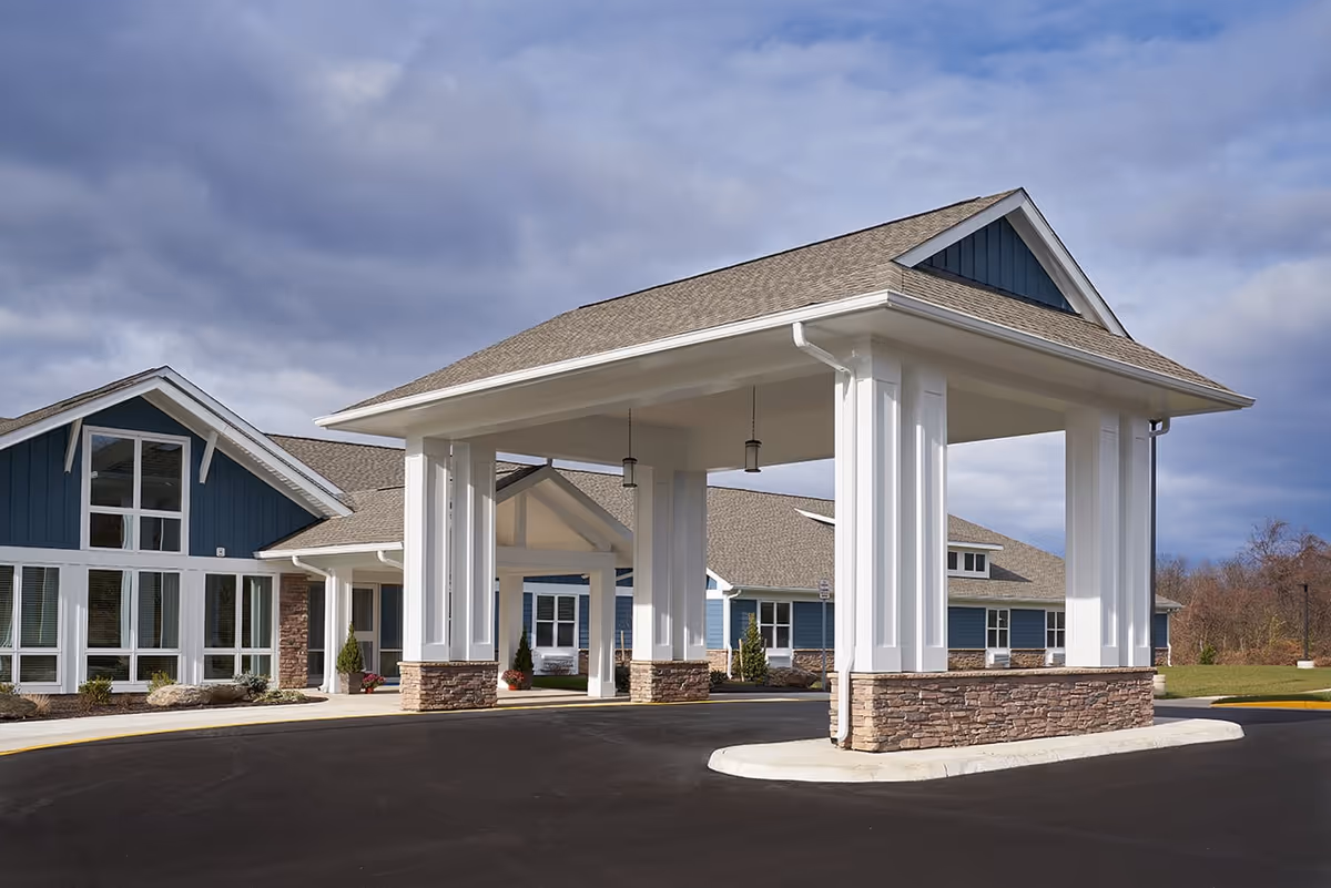 Exterior view of a senior living facility with a covered entrance supported by white columns with stone bases. The building has blue siding, large windows, and a sloped roof under a cloudy sky.
