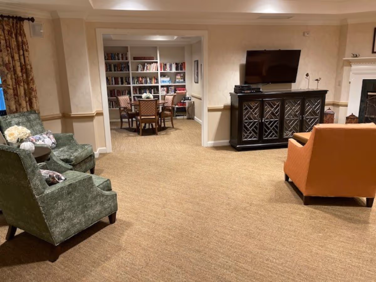 A cozy living room area with two green armchairs and an orange armchair arranged around a TV mounted on the wall above a dark wooden cabinet. In the background, there is a room with bookshelves filled with books and a round table with four chairs. The room has beige walls and carpeted floors.