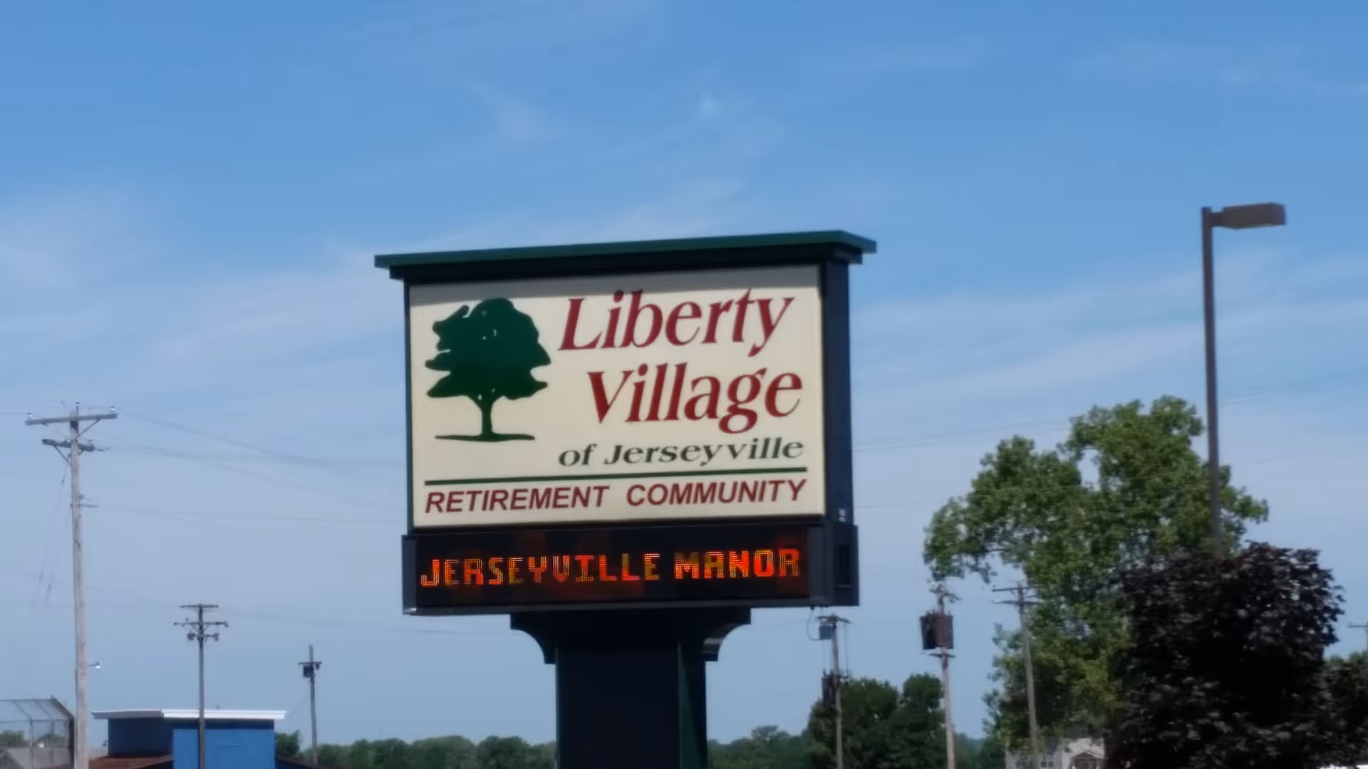 Outdoor sign for Liberty Village of Jerseyville Retirement Community with a green tree logo and an electronic message board displaying 'JERSEYVILLE MANOR'. The sign is set against a blue sky with some trees and utility poles in the background.