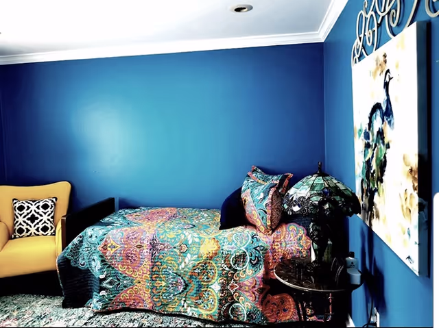 Small bedroom with a colorful patterned bedspread against a bright blue wall, a yellow armchair, decorative lamp, and wall art.