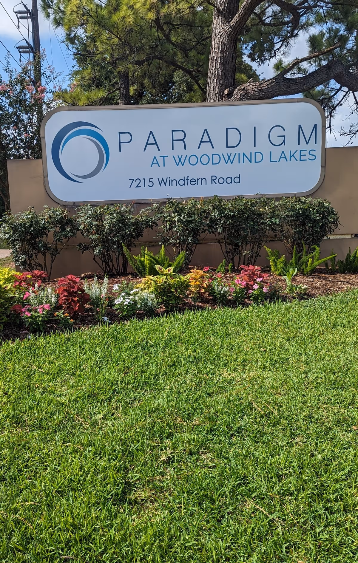 Entrance sign for Paradigm at Woodwind Lakes (7215 Windfern Road) above landscaped flowers and grass.