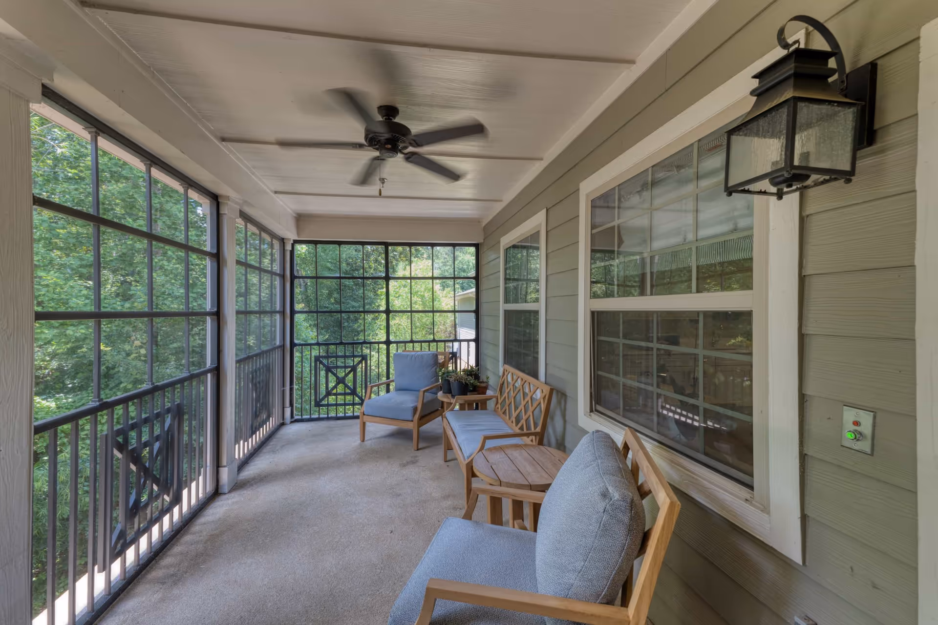 Screened porch with cushioned wooden chairs and small tables, a ceiling fan, and a view of surrounding trees.