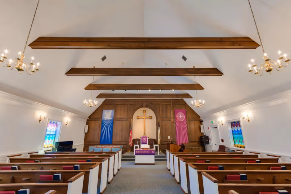 Interior of a chapel with wooden pews, stained glass windows, chandeliers, and a raised altar with a cross.