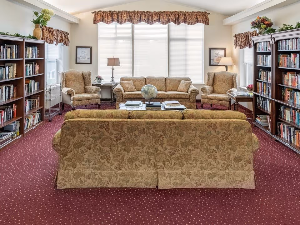 A cozy living room with patterned beige sofas and armchairs arranged around a coffee table with books and a globe. The room has red carpet flooring, large windows with blinds and valances, bookshelves filled with books on both sides, and table lamps providing warm lighting.