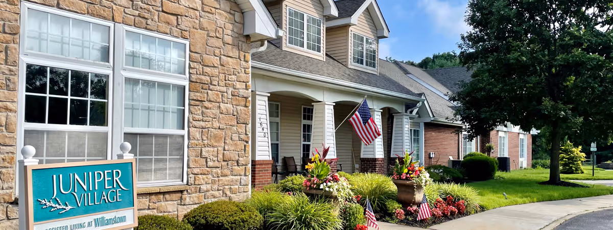 Exterior view of Juniper Village at Williamstown showing a stone and siding building with large windows, a porch with chairs, American flags, and a well-maintained garden with colorful flowers and shrubs along a curved sidewalk.