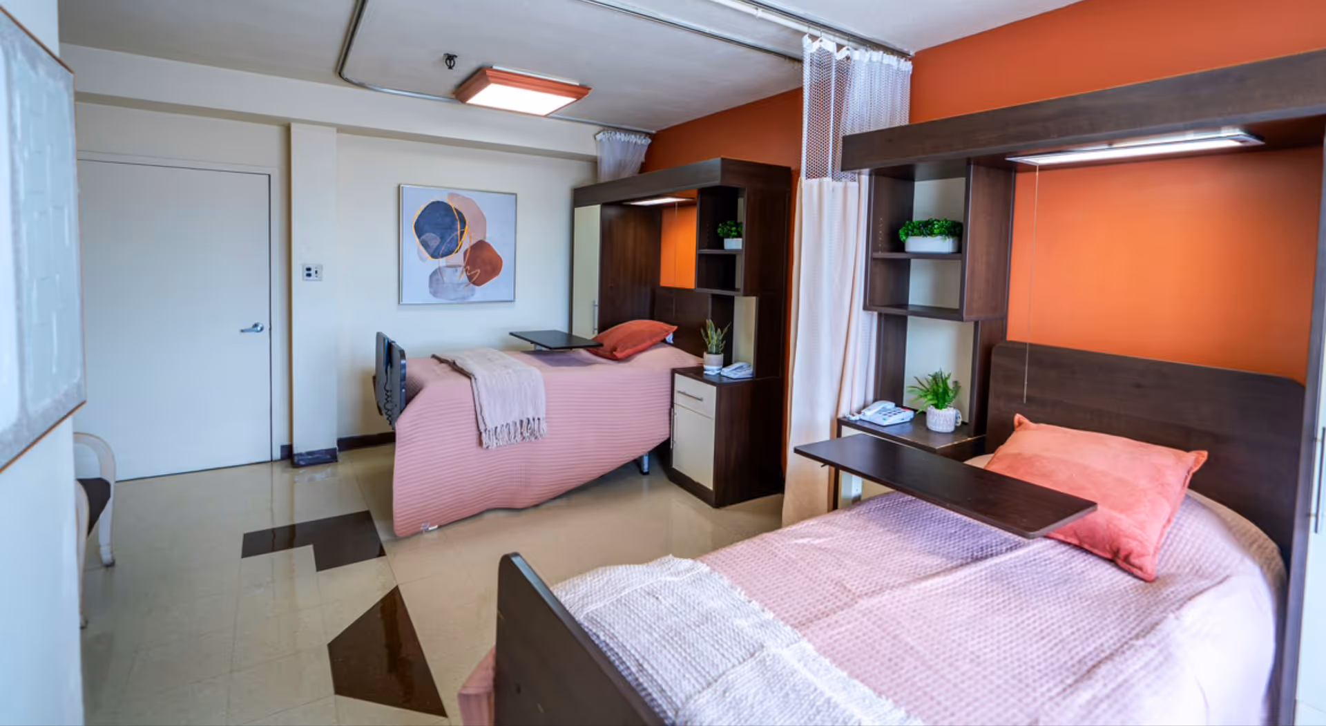 A nursing facility room with two single beds, each with a pink bedspread and a coral pillow. Each bed has a dark wooden headboard with built-in shelves holding small potted plants and a telephone. A curtain hangs between the beds for privacy. The walls are painted white and orange, and there is abstract artwork on the wall. The floor has a tiled pattern in beige and brown.