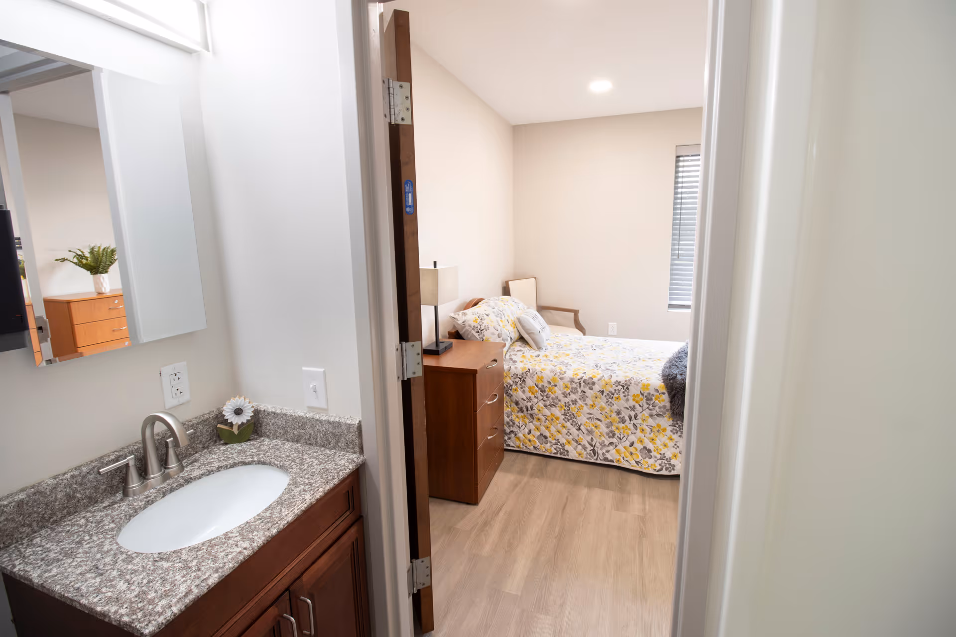 View through a doorway showing a small bedroom with a floral bedspread and a sink vanity in the foreground.