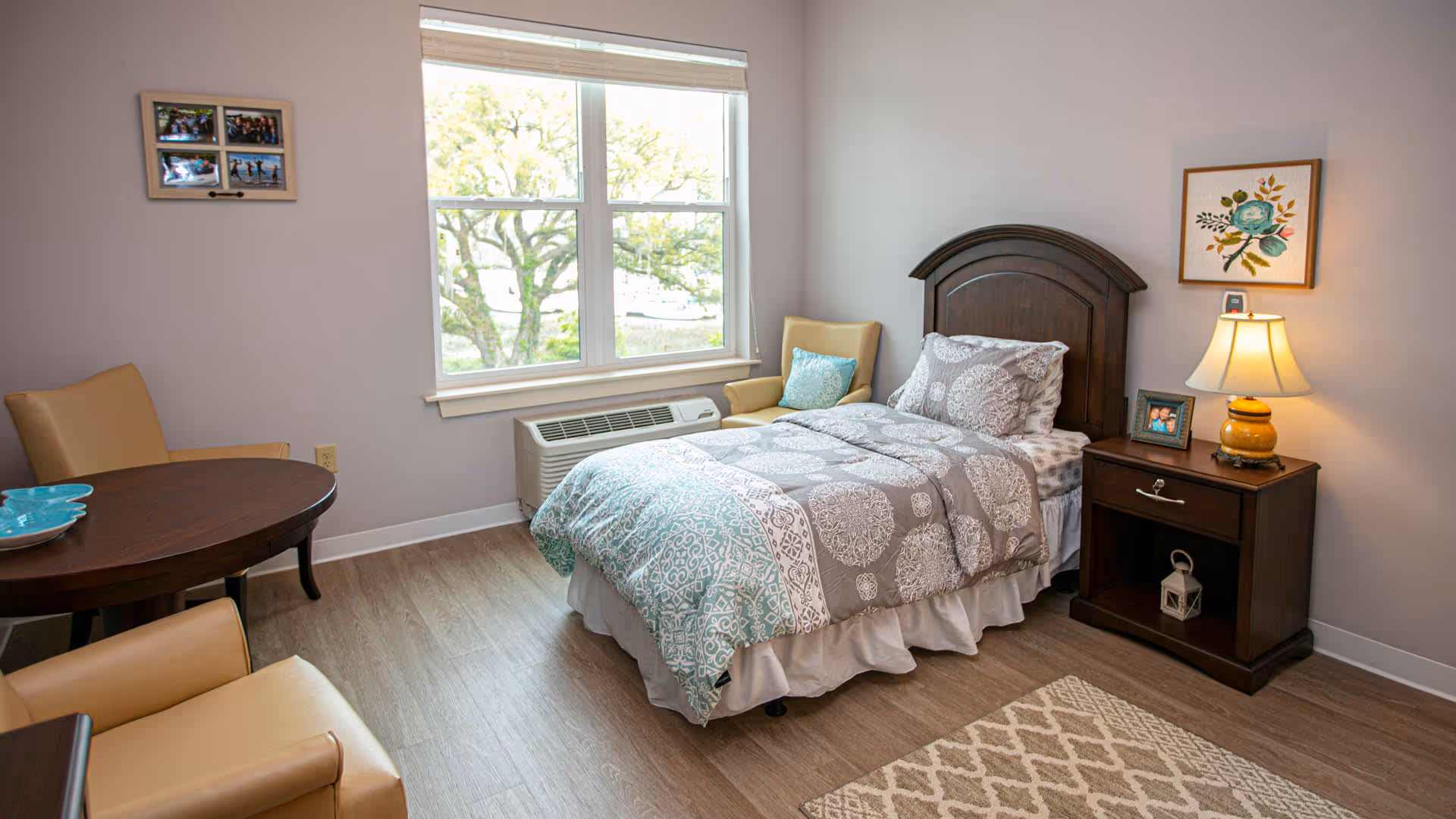 A cozy bedroom with a single bed featuring a patterned gray and teal comforter. Next to the bed is a wooden nightstand with a lamp, a framed photo, and a decorative item. There is a beige armchair with a blue cushion near a large window that lets in natural light. A round wooden table with beige chairs is partially visible in the foreground. The room has light-colored walls and wood flooring with a patterned rug.