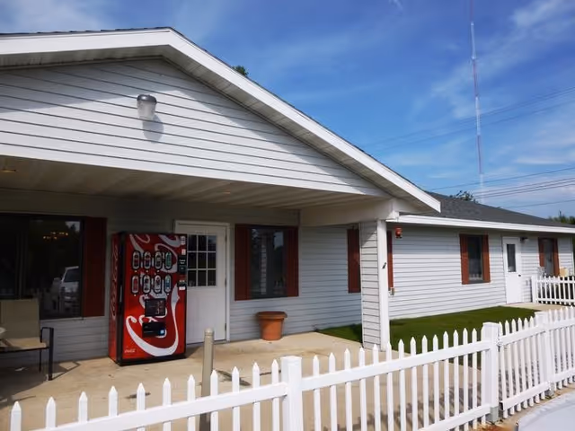 A single-story light gray building with a white picket fence, red shutters, and a vending machine.