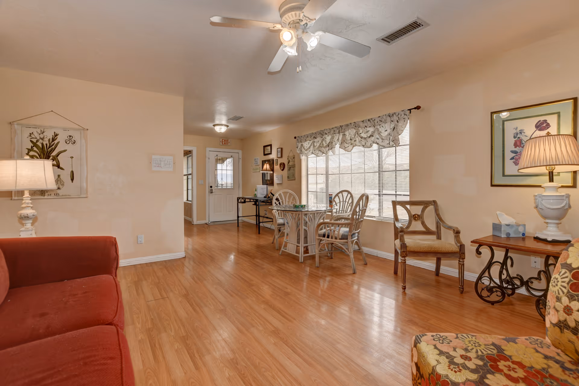 A bright and cozy living room area in Prescott Valley Assisted Living featuring a red sofa on the left, a small round table with four chairs near a large window with floral valance curtains, a wooden chair with armrests, a side table with a lamp and tissue box, and a ceiling fan with lights. The room has light-colored walls and wood flooring.