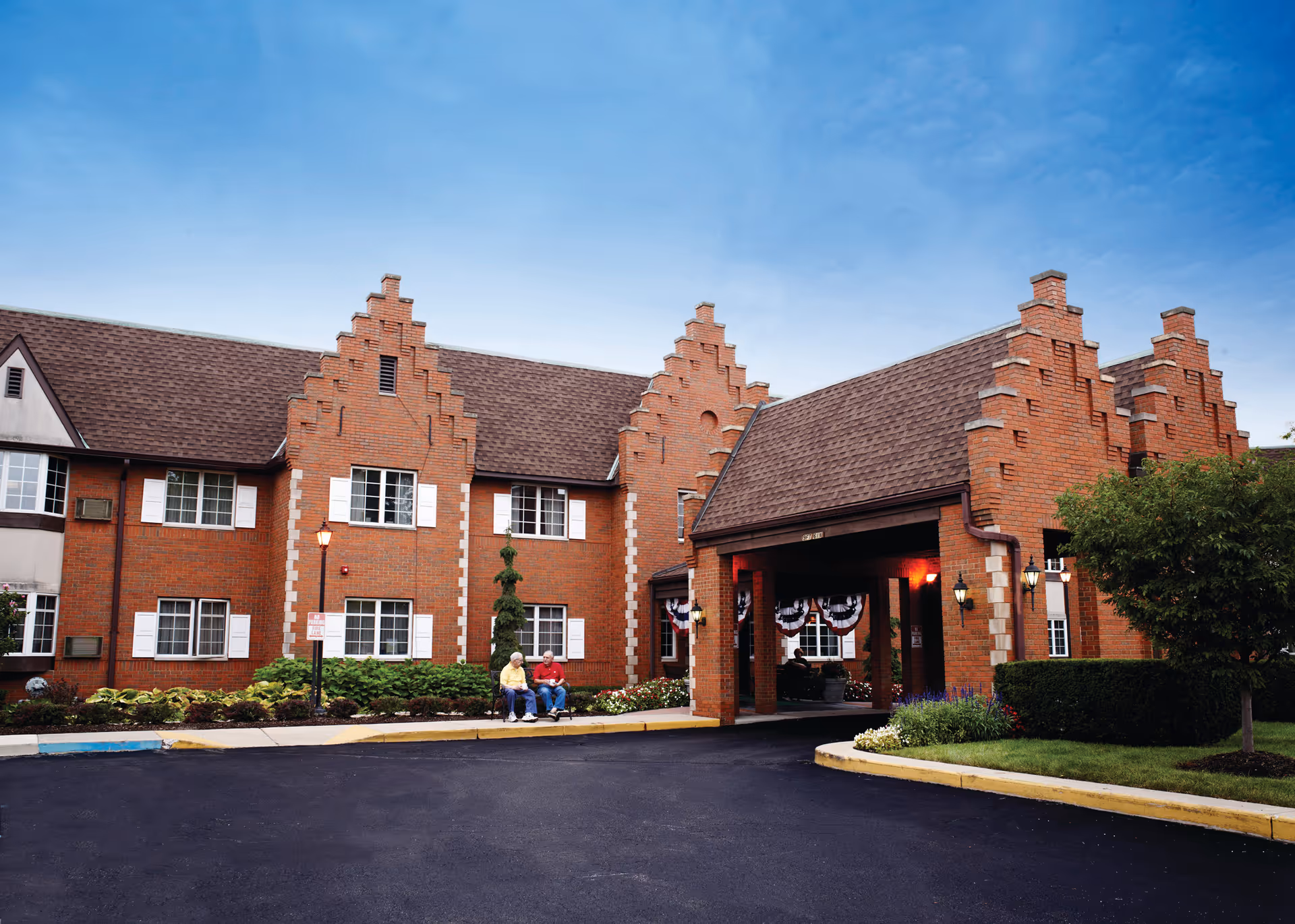 Exterior view of a brick senior living facility with a covered entrance. Two elderly people are sitting on a bench near the entrance. The building has a steep, stepped gable roof design and white window shutters. There are shrubs and a tree in the landscaped area near the entrance.