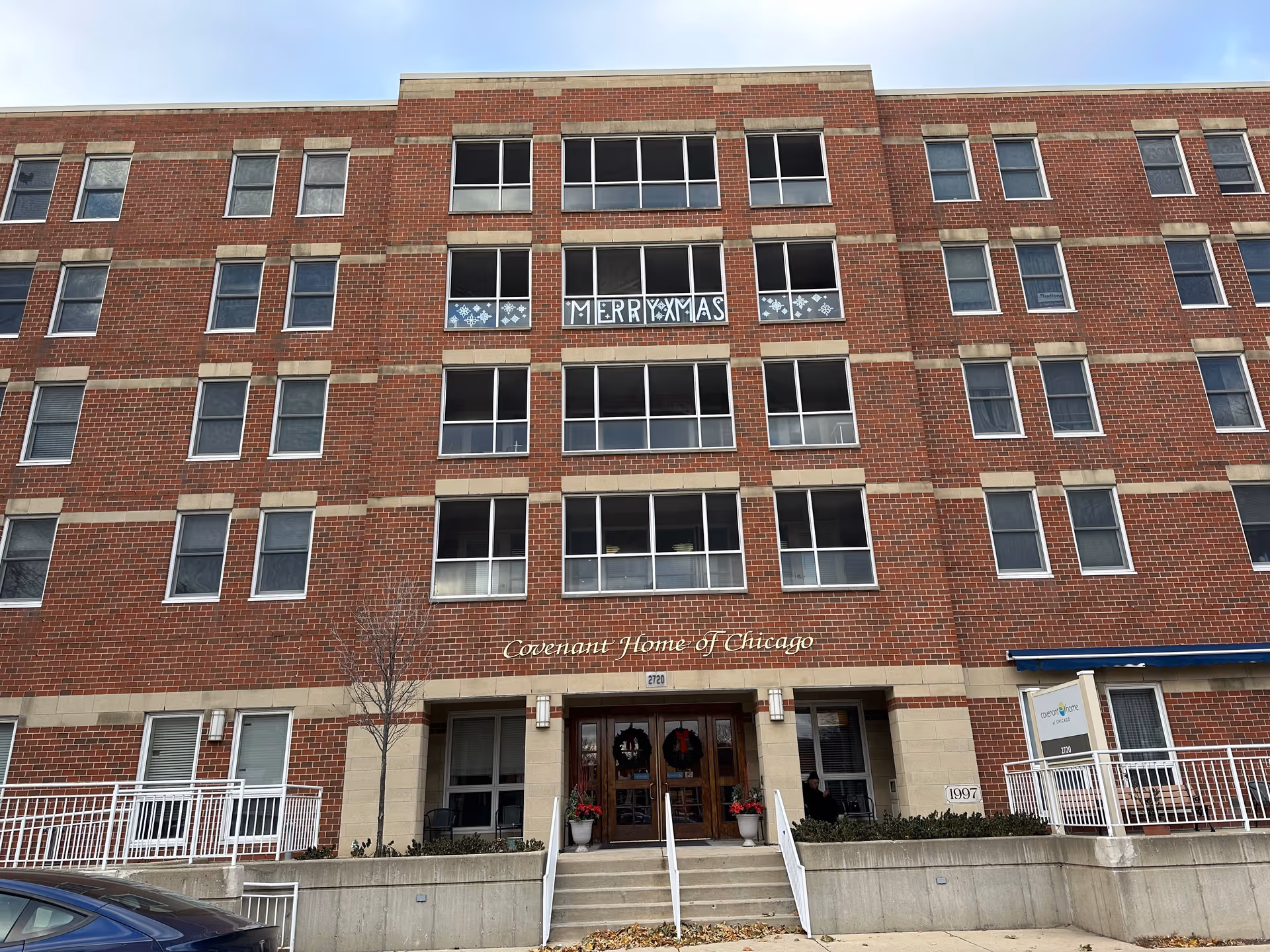 Front exterior view of a multi-story brick building named Covenant Home of Chicago. The building has multiple windows, some decorated with white snowflake patterns and a 'MERRY XMAS' sign. There are stairs leading up to double wooden doors adorned with holiday wreaths and red poinsettia plants. A blue car is partially visible in the foreground.