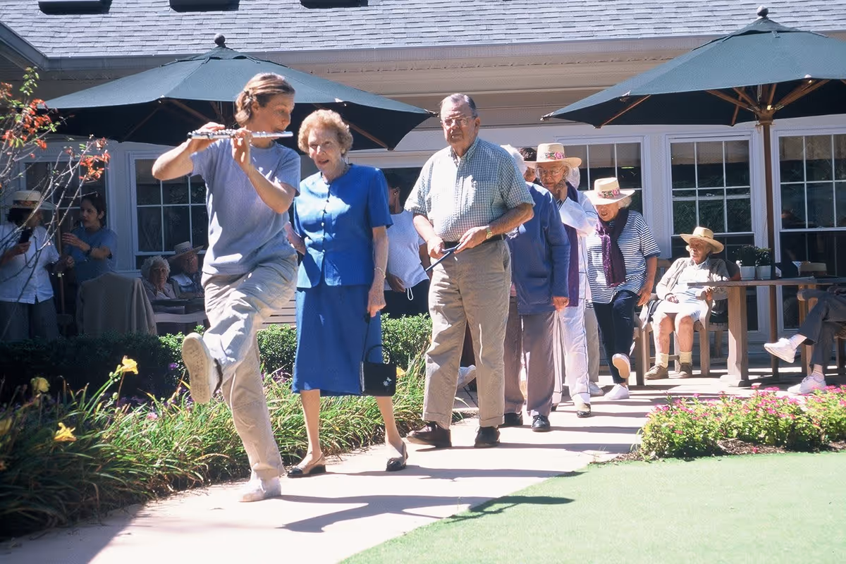 A group of elderly people walking in a line outdoors at a senior living facility, led by a younger person playing a flute. Some elderly individuals are seated under large green umbrellas near a building with large windows.