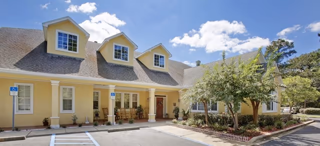 Exterior view of a yellow senior living facility building with a covered entrance, three dormer windows on the roof, and a small landscaped area with trees and shrubs in front. The sky is blue with some clouds.