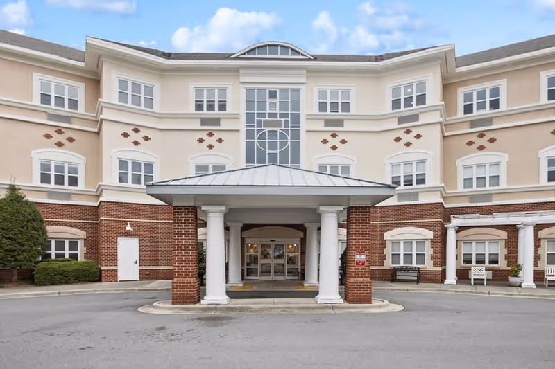 Front exterior view of a multi-story senior living facility building with a covered entrance supported by white columns, multiple windows, and a driveway in front. The building has a combination of beige and brick facade with decorative elements and some outdoor benches near the entrance.