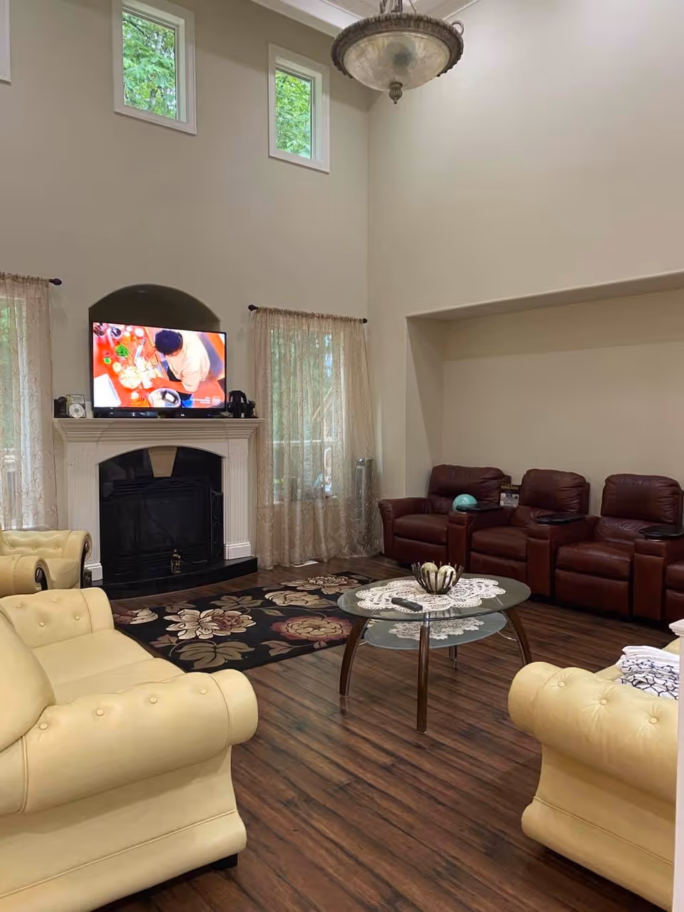 Spacious living room with high ceilings, a fireplace and TV above the mantel, cream sofas, brown leather recliners, and a glass coffee table on wood flooring.