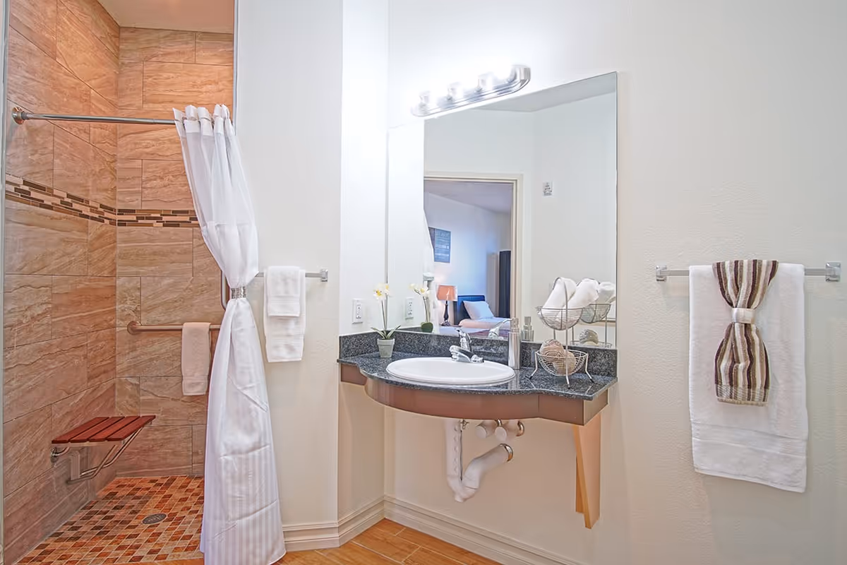 A clean and modern bathroom featuring a walk-in shower with brown and beige tiles, a white shower curtain tied back, a wall-mounted wooden shower seat, and a granite countertop with a white sink. The countertop holds decorative items including a small potted plant and a metal basket with rolled towels. A large mirror above the sink reflects part of a bedroom in the background. Towels hang on both sides of the sink area.