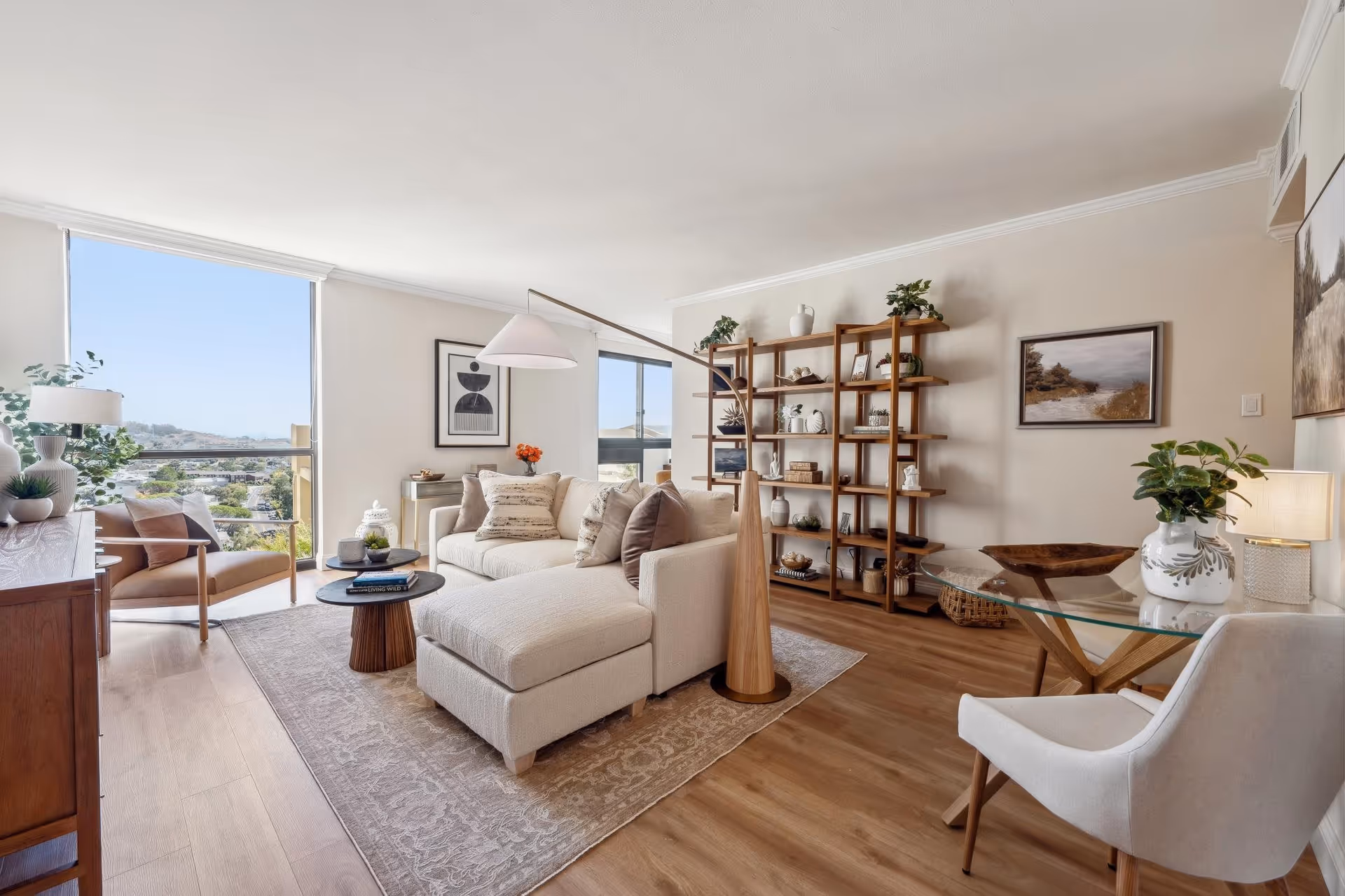 A bright and modern living room with large windows offering a view of the outdoors. The room features a beige sectional sofa with multiple cushions, a wooden coffee table with books and decorative items, a wooden shelving unit filled with plants and decor, a glass-top table with a white chair, and framed artwork on the walls. The floor is wooden with a patterned area rug under the sofa.