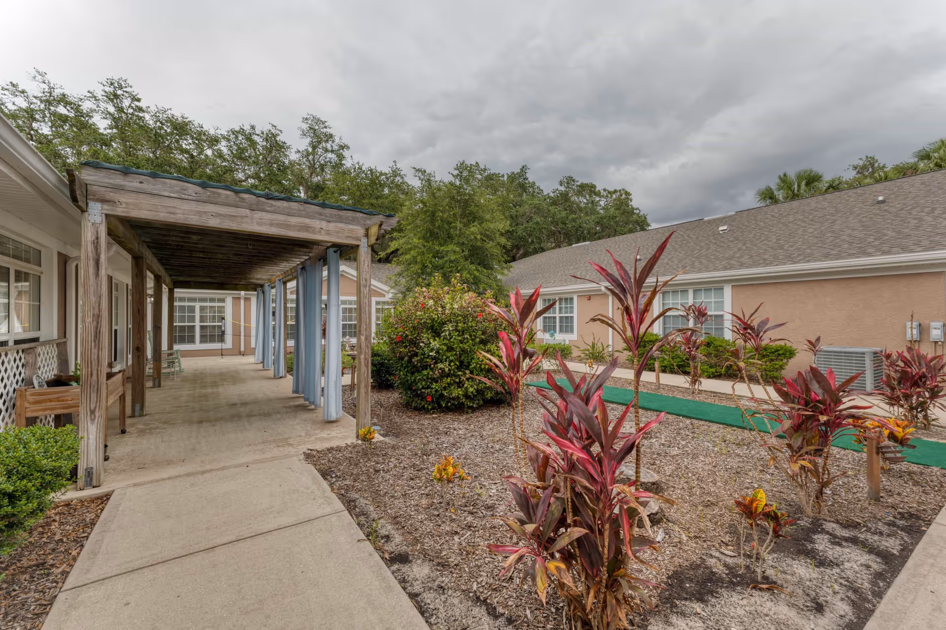 Outdoor courtyard area of a senior living facility with a wooden pergola on the left side, light blue curtains hanging from the pergola, a concrete walkway, landscaped garden beds with red and green plants, and beige buildings with white-trimmed windows in the background under a cloudy sky.