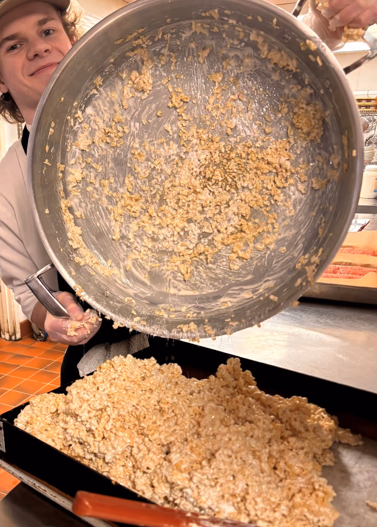 A person wearing a white shirt and gloves is pouring a large amount of a sticky rice cereal mixture from a big metal bowl onto a baking tray in a kitchen setting.