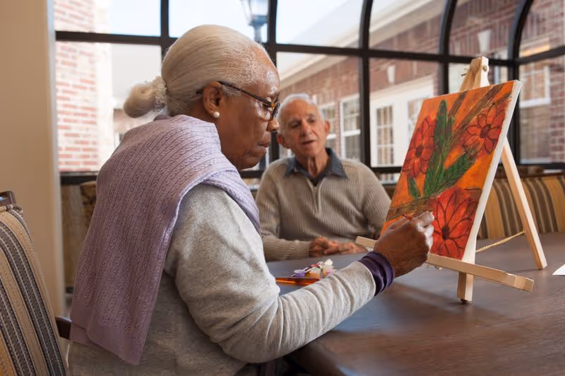 An elderly woman with gray hair tied back and wearing glasses and a lavender shawl is painting a colorful floral picture on an easel. An elderly man in a gray sweater sits nearby watching her. They are seated at a wooden table in a bright room with large windows and brick walls.