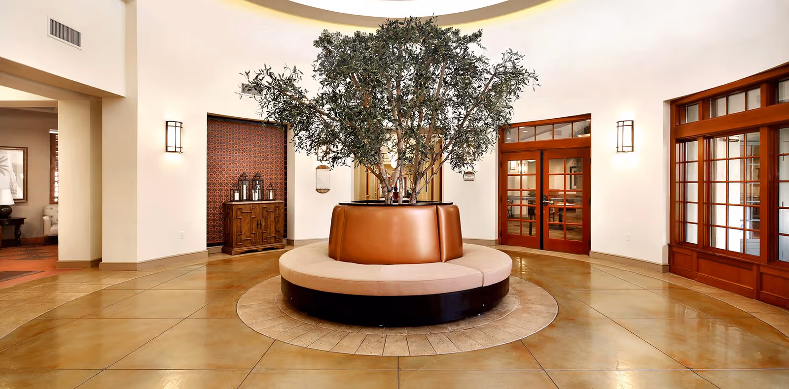 Bright circular lobby with a round bench surrounding a large potted indoor tree and wood-framed doors.