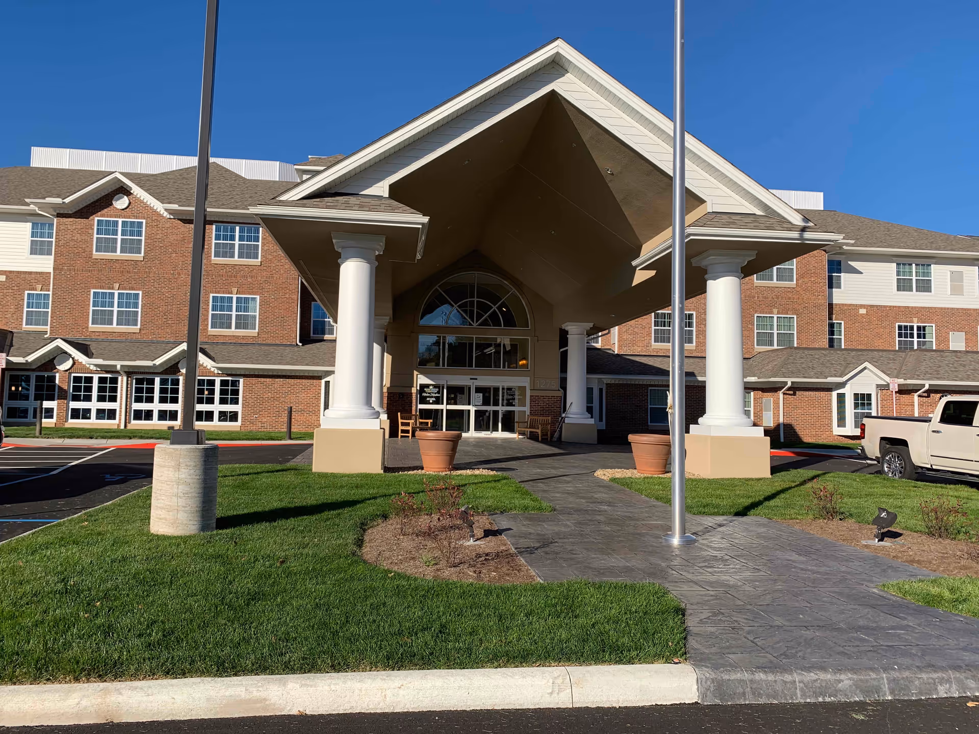 Front exterior view of Hudson Meadows Retirement Community building with a covered entrance supported by large white columns, a paved walkway, green grass, and a clear blue sky.