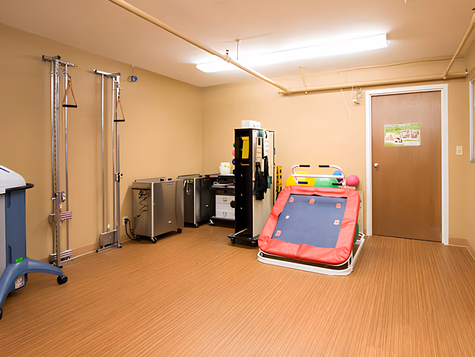 A therapy or rehabilitation room with beige walls and a brown door. The room contains exercise and therapy equipment including a red padded inclined therapy table, metal exercise apparatus with straps, and various storage carts. The floor has a wood-like pattern, and fluorescent lights are mounted on the ceiling.