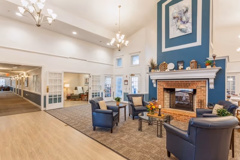 A bright and spacious assisted living facility common area with a high ceiling, featuring a brick fireplace with a blue accent wall above it. Four dark leather armchairs are arranged around a glass coffee table with a flower arrangement. The room has beige carpeting and light wood flooring, with chandeliers hanging from the ceiling. French doors and windows allow natural light to enter, and a hallway extends to the left.